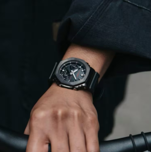 rimer plano de una persona usando el reloj G-SHOCK GM-2100CB-1A en la muñeca, mostrando su diseño moderno con bisel metálico negro y correa de tela.
